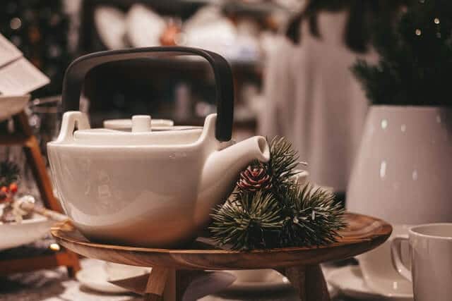 A white teapot with a sprig of spruce sat on a raised wooden plate on a laid table.