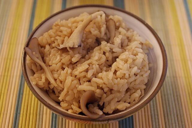 A small bowl on a striped tablecloth with cooked rice mixed with strips of maitake mushrooms.